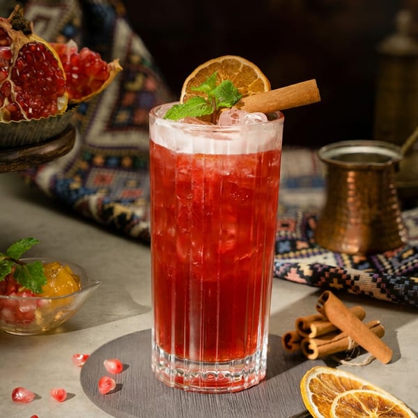 Vibrant red cocktail with dried orange, cinnamon, and mint, surrounded by pomegranates and a copper pot.